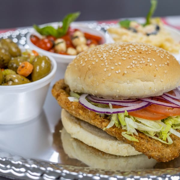 A Scimeca's Deli fried chicken sandwich with lettuce, tomato, and onion, served with sides.