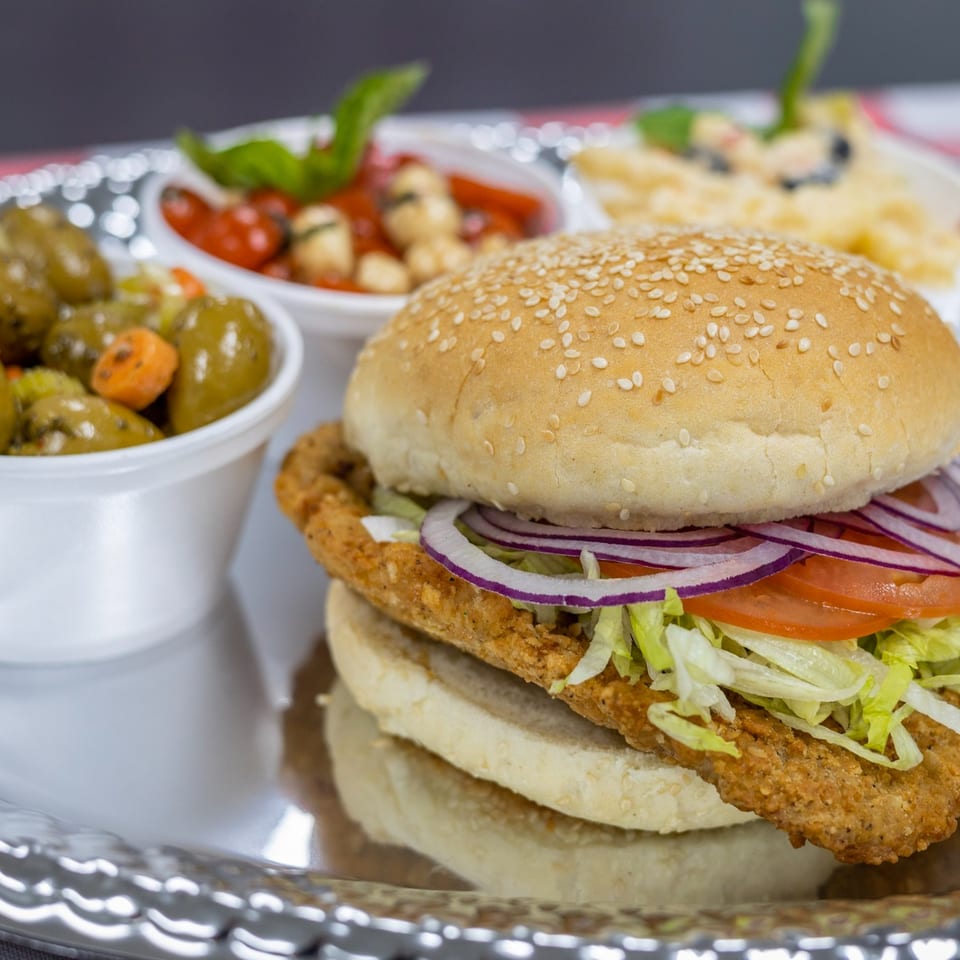 A Scimeca's Deli fried chicken sandwich with lettuce, tomato, and onion, served with sides.