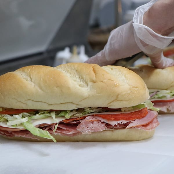 A gloved hand prepares a Scimeca's Deli sub sandwich with various meats, cheese, lettuce, and tomatoes.